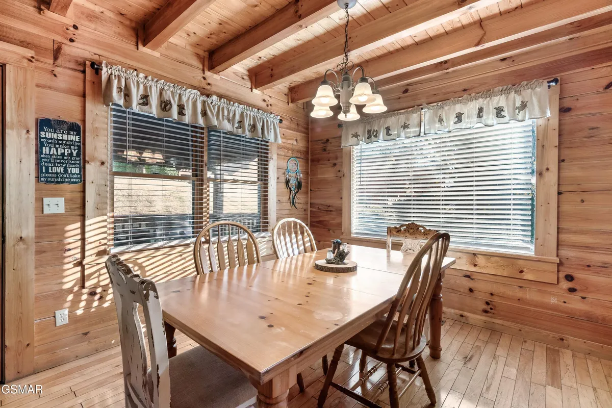 A bright and inviting dining area with a wooden dining table set for six, exposed wooden beams, and windows adorned with decorative curtains. The space exudes warmth and comfort, making it ideal for shared meals, morning coffee, or family board games.