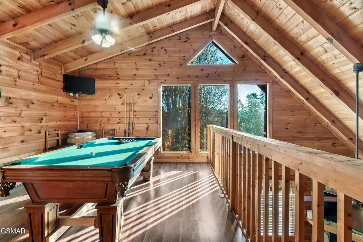 An interior shot of a bright game loft featuring a pool table, large windows, wooden beams, and comfortable seating. The space exudes a relaxed, playful atmosphere perfect for game nights and socializing while enjoying scenic views.