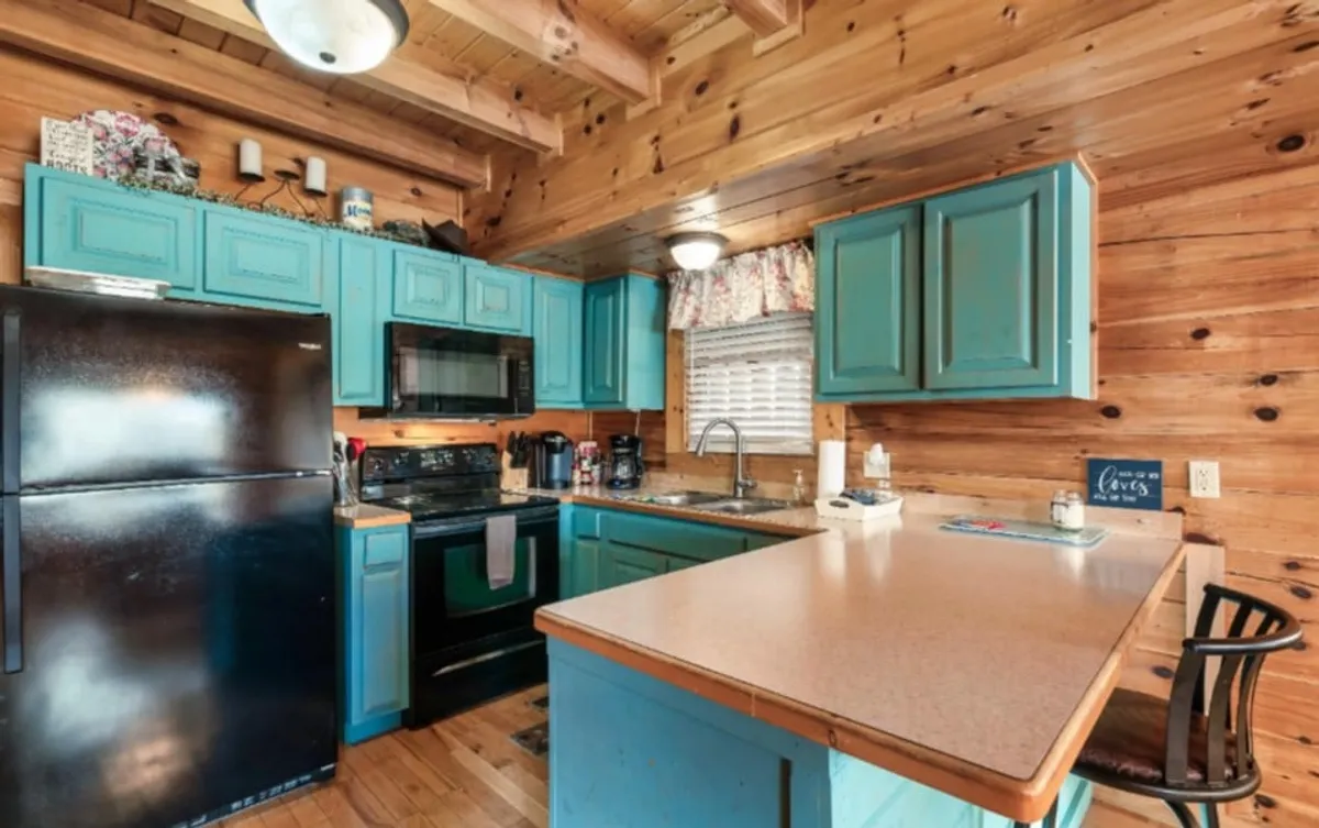 A cozy kitchen scene with turquoise cabinetry, black appliances, wooden accents, and convenient countertop seating. The open layout encourages both meal preparation and casual dining or conversation, reflecting the cabin’s social vibe.