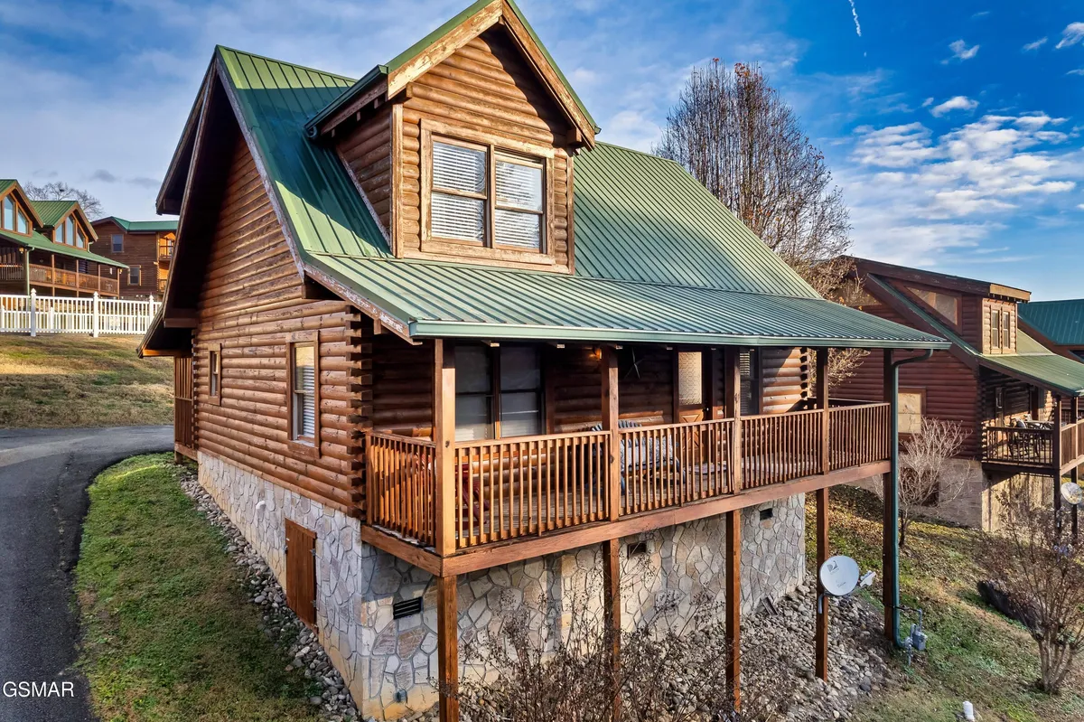 This exterior shot displays the charming log cabin structure with a green metal roof, a wooden porch, and natural surroundings. The porch invites guests to relax and enjoy the scenic environment, suggesting tranquility after a busy day out.