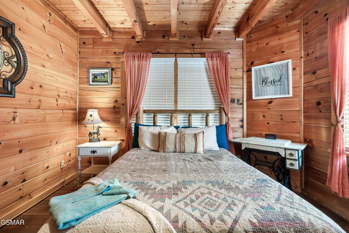 This image shows a welcoming bedroom with a bed adorned in a patterned quilt, flanked by wooden beams and a lamp on the nightstand. Framed artwork and curtains complete the inviting setup, creating a sense of warmth and comfort ideal for restful sleep.