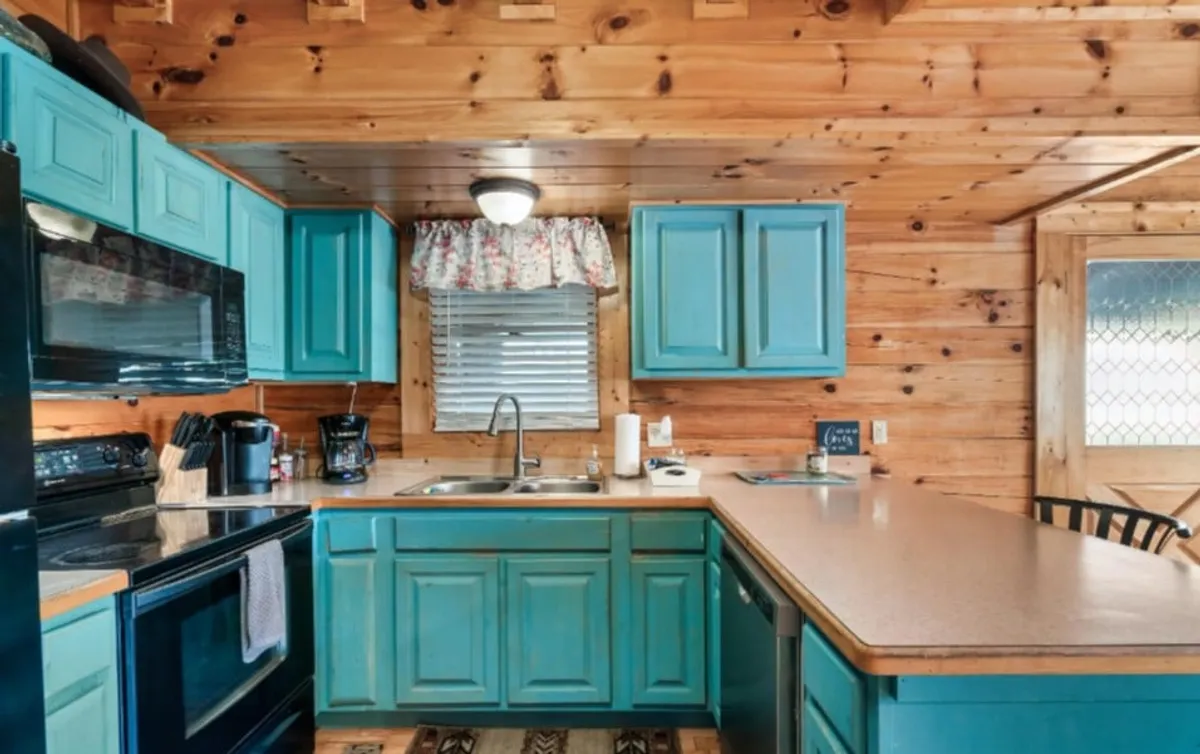 This image features a warmly lit kitchen with eye-catching turquoise cabinets, black modern appliances, wooden countertops, and a colorful floral curtain. The space feels inviting and practical, blending rustic charm with updated essentials ideal for preparing family meals.