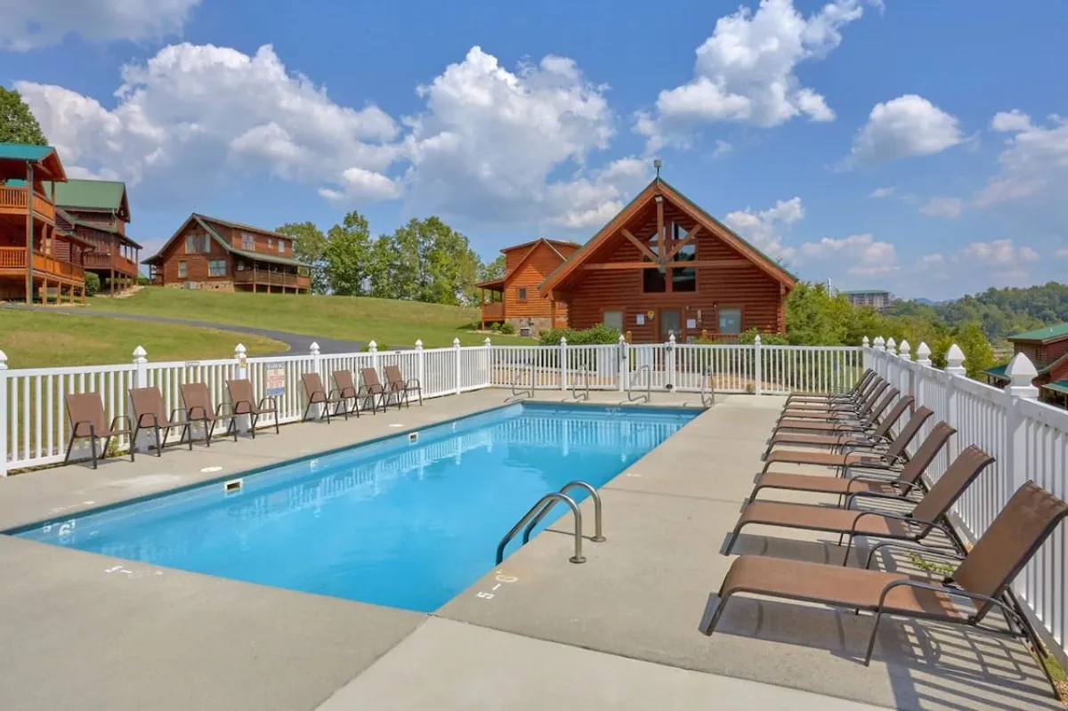 This exterior shot centers on a sparkling swimming pool surrounded by lounge chairs and classic log cabin architecture in the background. The atmosphere is one of laid-back leisure, perfect for sunbathing, swimming, or social gatherings.