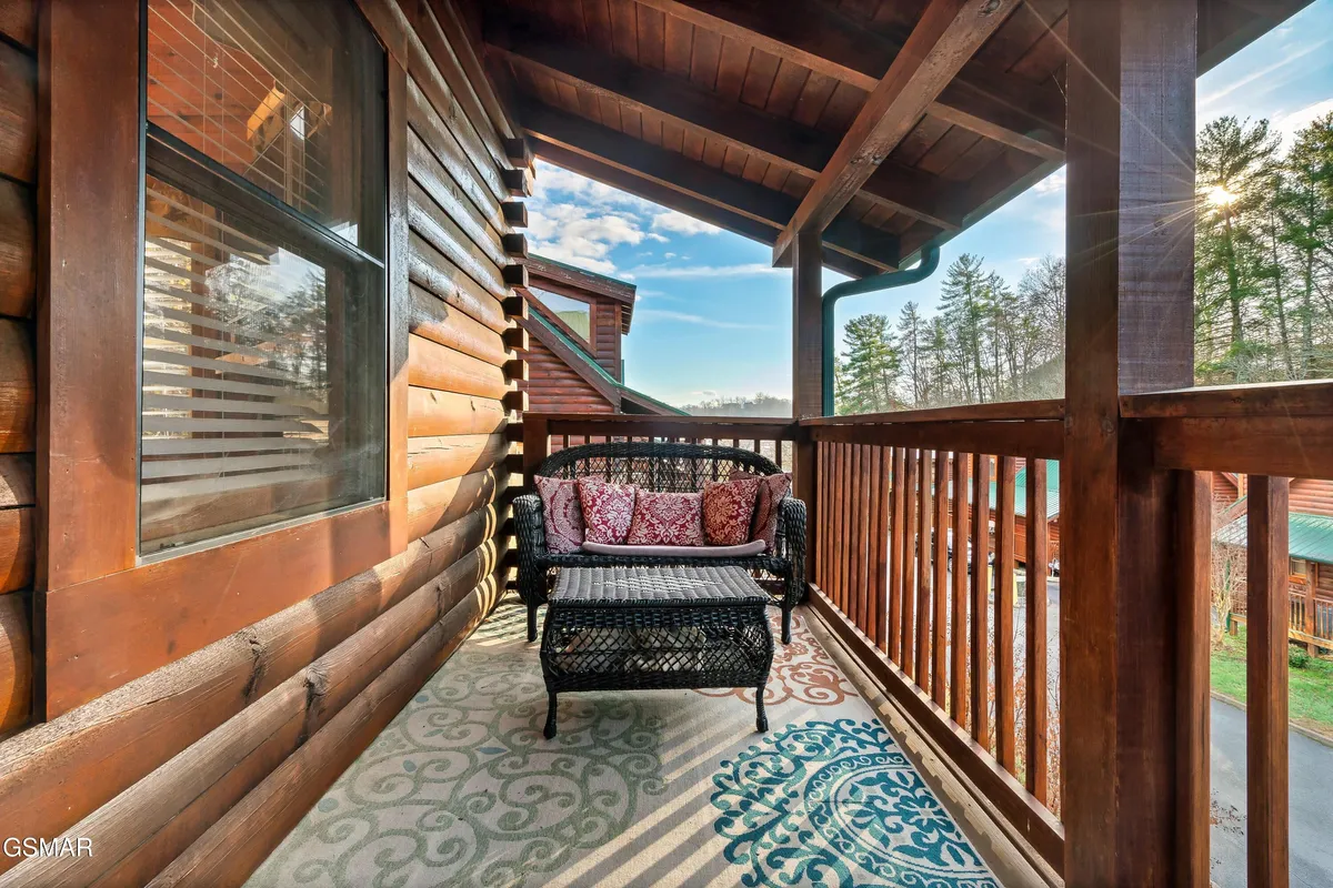 An inviting outdoor seating area with a wicker chair, decorative pillows, a patterned rug, and a wooden railing. Surrounded by nature, it's a cozy spot for relaxing, reading, or socializing while taking in the outdoors.