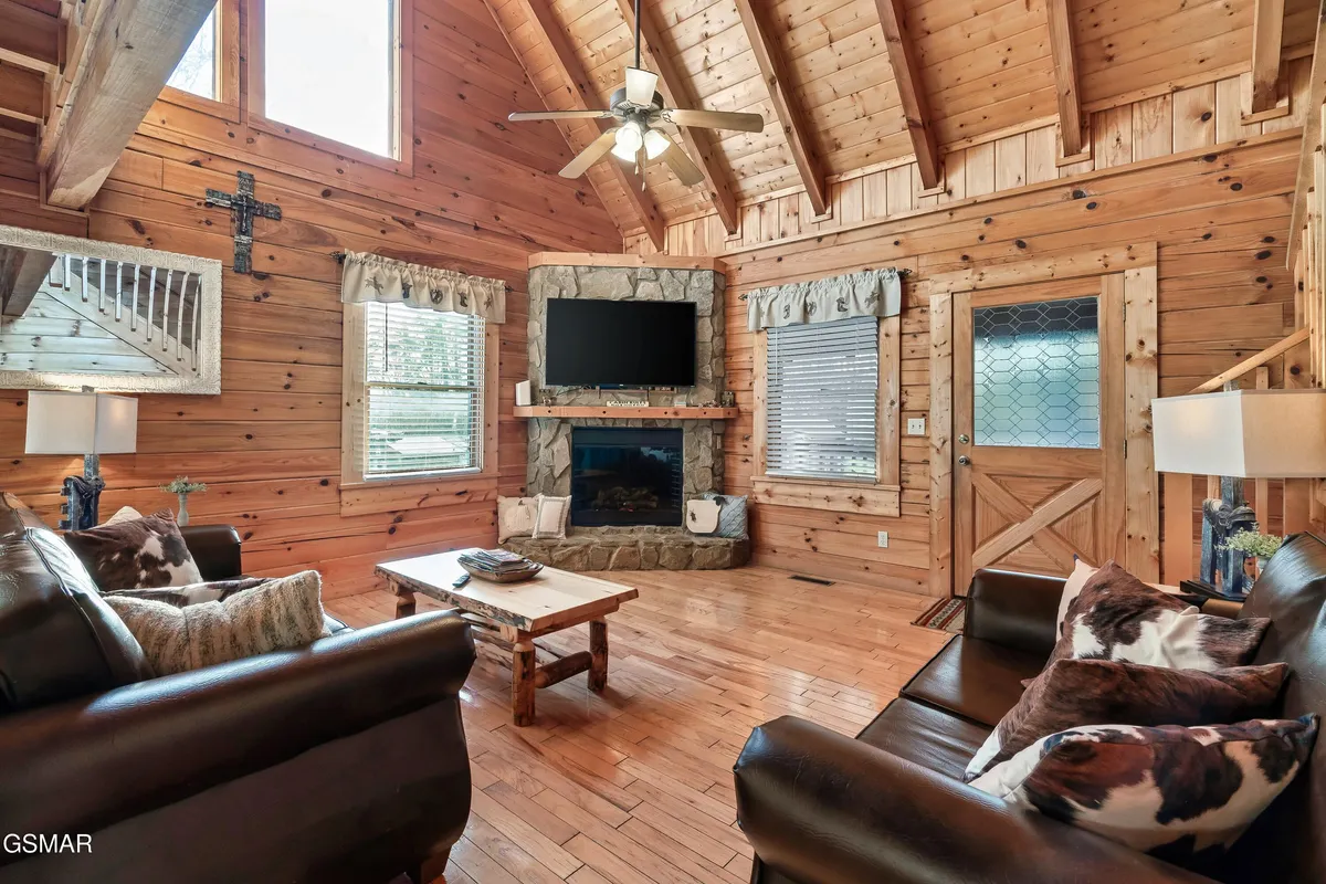 This interior image showcases a spacious living room with a striking stone fireplace, wooden ceiling, large windows, and an arrangement of comfortable seating. The inviting setting emphasizes cozy gatherings, relaxation, and enjoying the warmth of the fire.