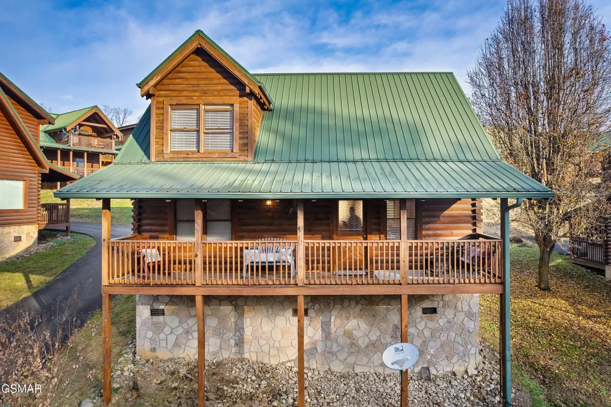 The image shows the inviting exterior of a classic log cabin with a green metal roof and a welcoming front porch furnished with comfortable seating. Surrounded by lush greenery, the porch overlooks the natural landscape, establishing a strong connection to the outdoors and the adventure that awaits nearby.